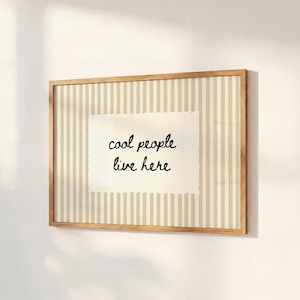 May include: A framed wall art piece with a light brown wooden frame. The artwork features a cream-colored background with vertical beige stripes. A white rectangular paper with the text "cool people live here" is centered.