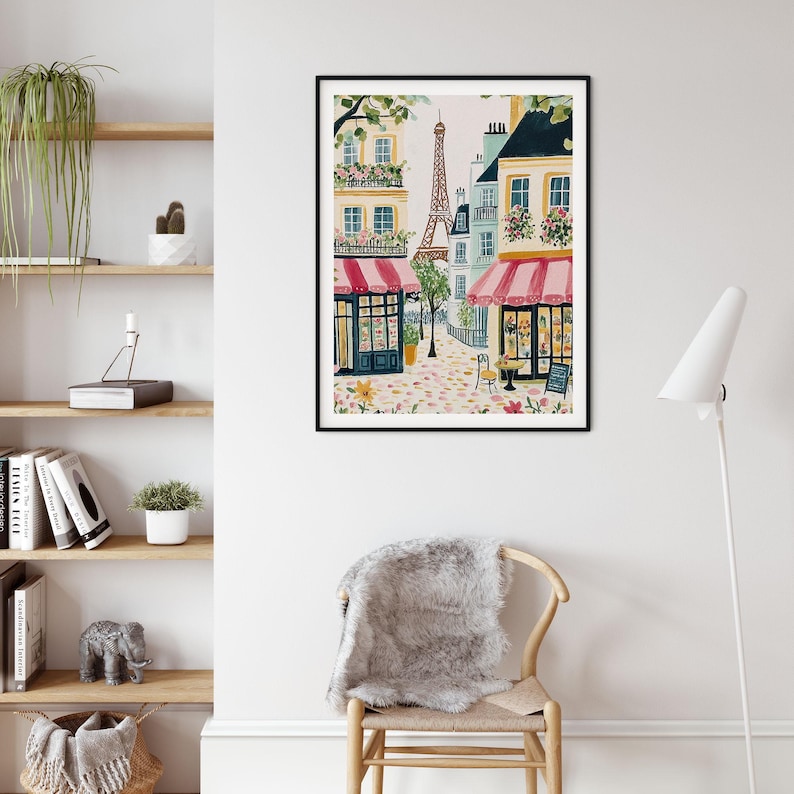 May include: Watercolor painting of a Parisian street scene with the Eiffel Tower in the background. Buildings with pink awnings and flower boxes line the cobblestone street. A black framed art print adds a touch of Parisian charm to any space.
