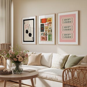 May include: Three framed art prints. The left print features cherries and the text "cherish the day". The middle print has various vintage-style graphics. The right print displays "LUCKY STRIKE" tickets on a pink background. The prints are displayed above a white sofa.