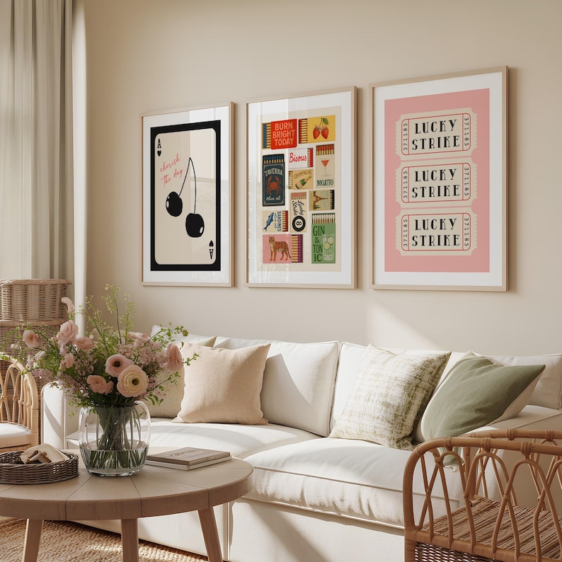 May include: Three framed art prints. The left print features cherries and the text "cherish the day". The middle print has various vintage-style graphics. The right print displays "LUCKY STRIKE" tickets on a pink background. The prints are displayed above a white sofa.