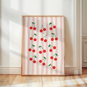 Könnte beinhalten: Ein gerahmter Kunstdruck mit einem Muster aus roten Kirschen mit grünen Stielen auf einem Hintergrund aus vertikalen rosa und cremefarbenen Streifen. Der Holzrahmen unterstreicht das fröhliche Design des Kunstwerks, ideal für Küchen- oder Esszimmerdekoration.