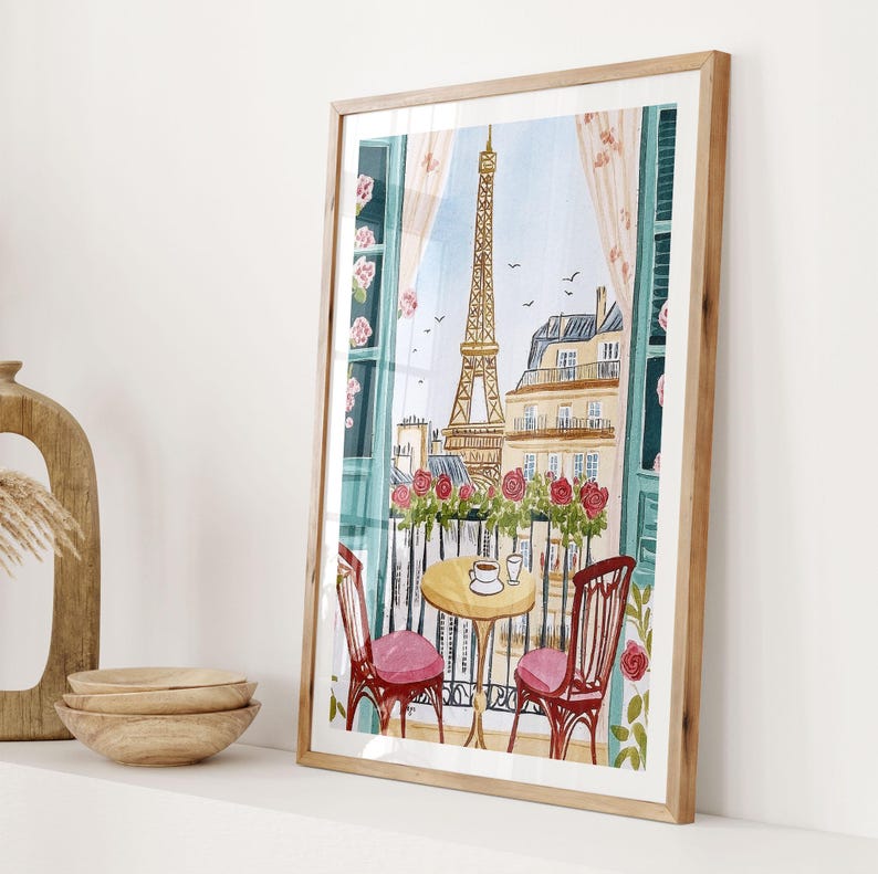 May include: Watercolor print of a Parisian balcony scene with the Eiffel Tower in the background. The artwork features a small table with two chairs, a cup of coffee, and a view of the city. The frame is a light wood color.