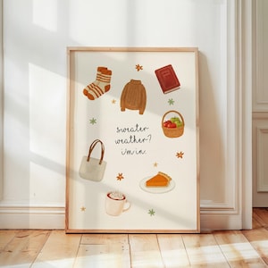 May include: A framed print with a cream background features illustrations of fall items. A brown sweater, socks, a book, a basket of apples, a purse, a slice of pie, and a mug of cocoa are displayed. The text reads "sweater weather? i'm in."