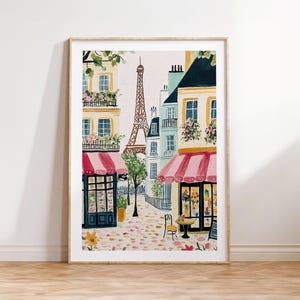 May include: Watercolor painting of a Parisian street scene with the Eiffel Tower in the background. Buildings with pink awnings and flower boxes line the street. A small table and chairs are set up outside a shop. The artwork is framed in a light wood frame.