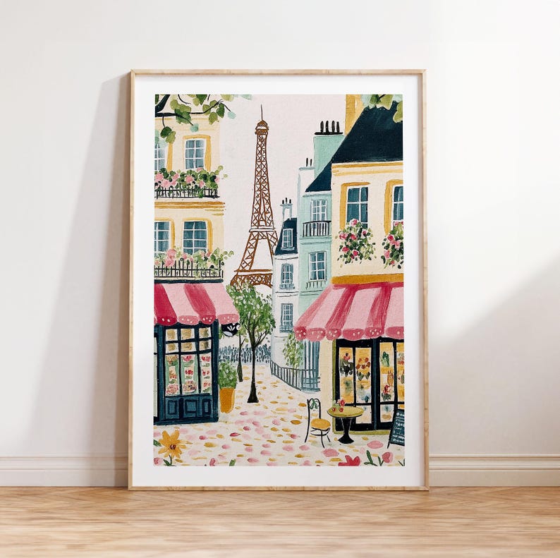 May include: Watercolor painting of a Parisian street scene with the Eiffel Tower in the background. Buildings with pink awnings and flower boxes line the street. A small table and chairs are set up outside a shop. The artwork is framed in a light wood frame.
