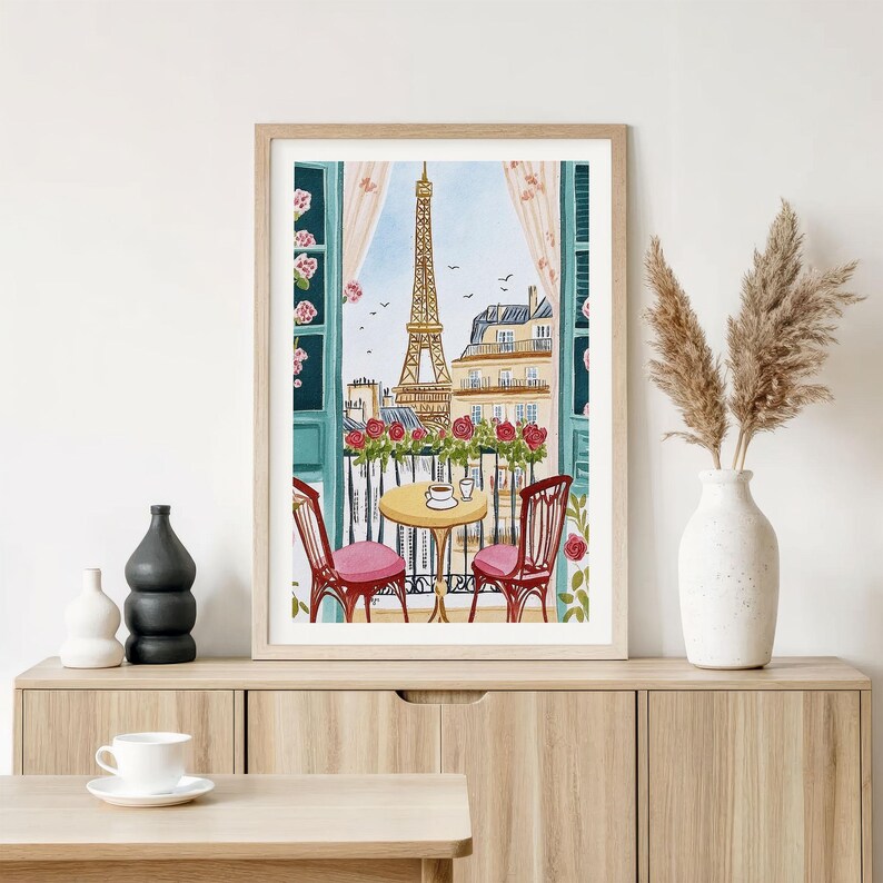 May include: Watercolor print of a Parisian balcony scene. The Eiffel Tower is visible through an open window with red roses, a small table, and two chairs. The artwork is framed in a light wood frame.