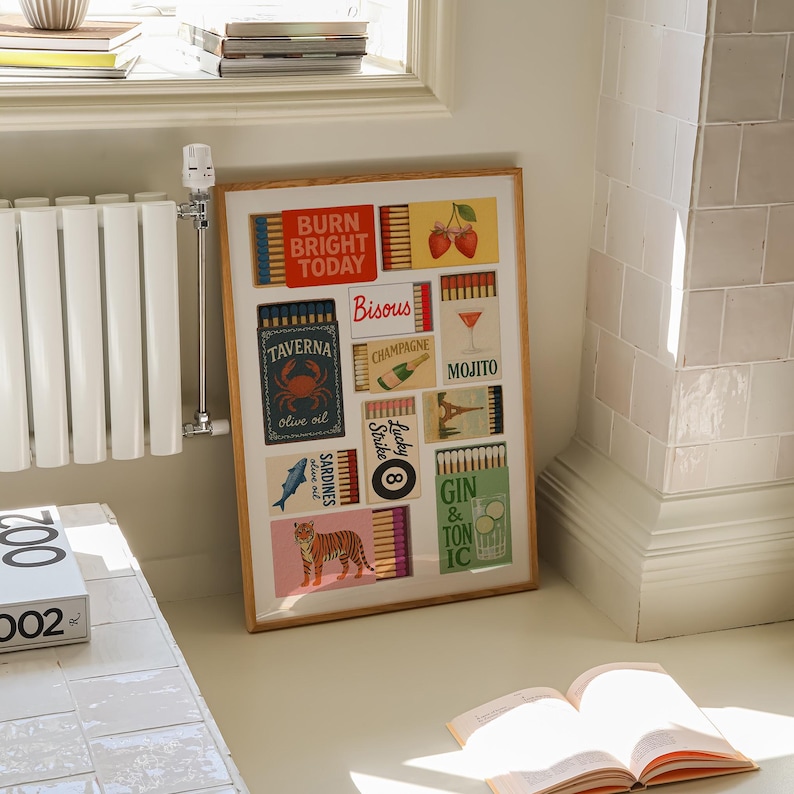May include: A framed artwork featuring colorful matchbox designs with various themes. The artwork includes text such as "BURN BRIGHT TODAY", "TAVERNA", "Bisous", "Champagne", "Mojito", "Lucky Strike", and "Gin & Tonic". The artwork is displayed on a white wall.