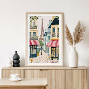 May include: Watercolor print of a Parisian street scene featuring the Eiffel Tower, shops with red awnings, and cobblestone streets. The artwork is framed in a light wood frame and displayed on a wooden cabinet.