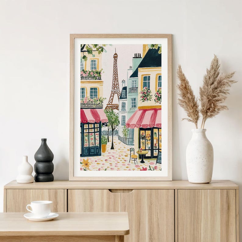 May include: Watercolor print of a Parisian street scene featuring the Eiffel Tower, shops with red awnings, and cobblestone streets. The artwork is framed in a light wood frame and displayed on a wooden cabinet.