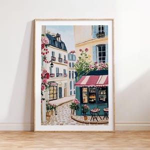 Könnte beinhalten: Ein Aquarellgemälde einer europäischen Straßenszene mit Kopfsteinpflaster, mehrstöckigen Gebäuden mit blauen Fensterläden und einem Ladenlokal mit einer rosa-weiß gestreiften Markise. Rosa Blumen ranken an den Wänden und schmücken die Cafétische.