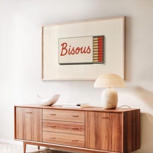 May include: A framed print of a white matchbox with the word "Bisous" in red cursive lettering. The matchbox is open and contains red and white matches.