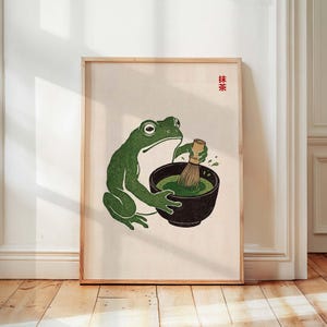 Pode incluir: Uma impressão artística emoldurada com uma ilustração de um sapo verde preparando chá matcha em uma tigela preta. O sapo é retratado em um estilo japonês. Os caracteres japoneses para "Matcha" estão no canto superior direito.