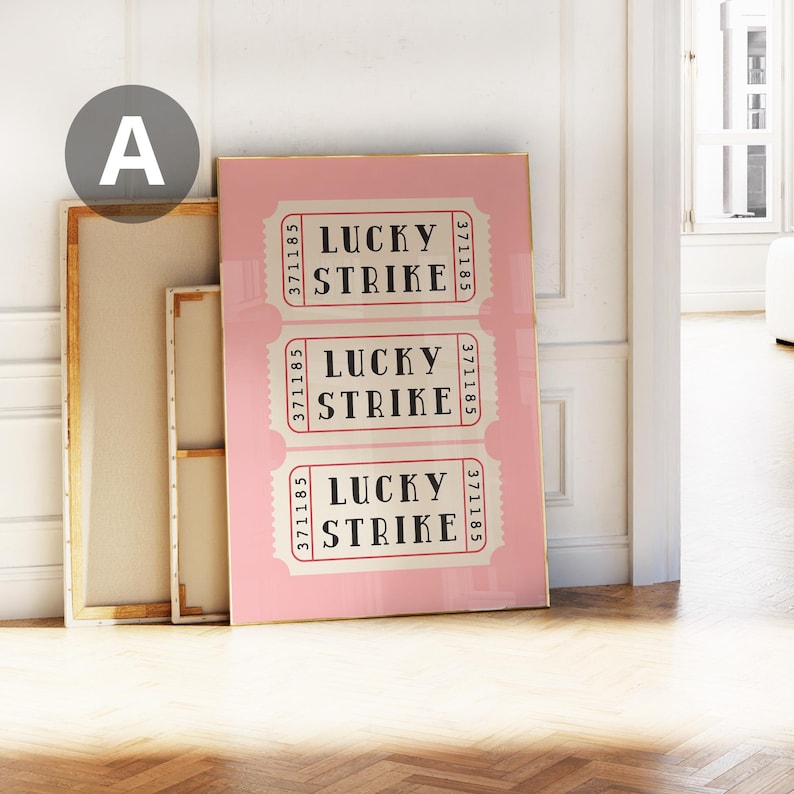 May include: A pink framed print featuring three vintage-style Lucky Strike ticket designs. Each ticket has the text "LUCKY STRIKE" in a bold font, with serial numbers. The print is leaning against a wall with other framed art.