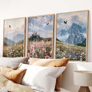 May include: A set of three framed landscape paintings featuring a castle on a hill, snow-capped mountains, and fields of pink and purple wildflowers under a cloudy sky with birds flying in the distance, displayed in light wooden frames.