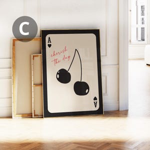 May include: A framed art print featuring a playing card design with a black border. The card displays two black cherries and the words "cherish the day" in red script. The Ace of Hearts and Ace of Spades symbols are also visible.