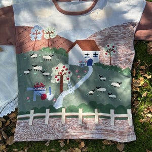Puede incluir: Un suéter de punto con una escena de paisaje pastoral que muestra una casa blanca, un camino sinuoso, una valla blanca y varias ovejas pastando en un campo verde. El diseño incluye árboles con frutos rojos, una carretilla y mangas marrones.