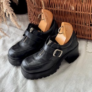 May include: A pair of black leather platform shoes with a zip and buckle fastening. The shoes have a chunky sole and a rounded toe. The shoes are displayed with wooden shoe trees inside.