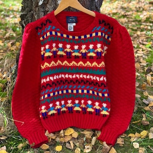 Puede incluir: Un suéter rojo con cuello redondo y mangas largas. El suéter presenta un patrón colorido de figuras con vestidos, con detalles en azul, amarillo, verde y blanco. El suéter está colgado de una percha de madera.