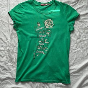 Puede incluir: Una camiseta verde con cuello redondo y mangas cortas. La camiseta presenta un gráfico de una rosa y el texto "The Wonderful World of Juicy Couture" en un diseño de banner. La camiseta está hecha de un material suave.