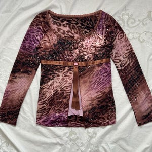 May include: A long-sleeved top with a brown, purple, and pink animal print design. The top features a scoop neckline, a brown ribbon belt with a button, and a light pink panel down the front. Small sequins are scattered across the fabric.
