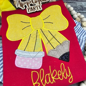 May include: A red shirt with a yellow bow and two pencils tied to the bow. The pencils are gray and brown with a pink eraser. The text "Applique Party" and "Blakeely" are embroidered on the shirt.