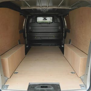 Könnte beinhalten: Innenansicht eines Transporters mit Sperrholzverkleidung und -boden. Das Innere des Transporters ist mit hellen Holzpaneelen ausgekleidet, einschließlich Wänden, Boden und eingebauten Staukästen. Die Hecktüren sind geöffnet.