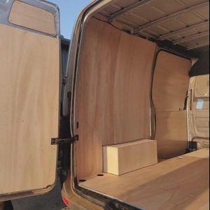 May include: Interior view of a van with plywood paneling and a rectangular wooden box. The van's side door is open, revealing the light-colored wood. The interior is unfinished, with the wood panels covering the walls and floor.