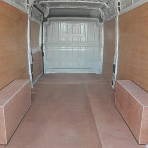 May include: Interior view of a white van with wooden paneling and flooring. The van's interior features light brown wood paneling on the walls and floor, with two rectangular wooden boxes on the floor. The van's back doors are open.