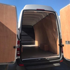 May include: A silver cargo van with open rear doors, showcasing a large interior. The inside is lined with light-coloured wooden panels. A small wooden box is visible inside the van.