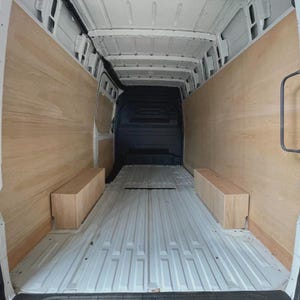 May include: Interior view of a cargo van with light wood paneling on the walls and two rectangular wooden boxes. The floor is a ribbed, light gray metal. The back of the van is black.