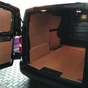Op de afbeelding: Zwarte bestelwagen met geopende achterdeuren, die een interieur van multiplex onthullen. De vloer en zijpanelen zijn gemaakt van lichtbruin multiplex, wat een schone en georganiseerde ruimte biedt. De buitenkant van de bestelwagen is zwart.