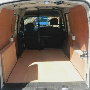 Può includere: Vista interna di un furgone bianco con pavimento e pannelli laterali in legno. Una gabbia metallica nera separa i sedili anteriori dall'area di carico. Il furgone è vuoto, con una vista dell'esterno visibile attraverso il finestrino posteriore.