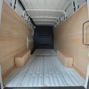 May include: Interior view of a cargo van with wooden paneling on the walls and two rectangular wooden boxes. The floor is a light gray, ribbed metal. The van's interior is empty and ready for customization.