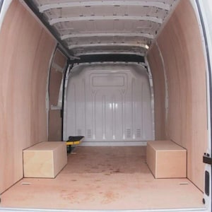Puede incluir: Vista interior de una furgoneta blanca con paneles de madera en las paredes y el suelo. Dos cajas rectangulares de madera están colocadas en el suelo. Las puertas traseras de la furgoneta están abiertas.