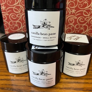 May include: Three small glass jars with black lids and white labels. The labels have a black and white illustration of a vanilla bean and the text "vanilla bean paste" and "HANDMADE - SMALL BATCH 1 TSP PASTE = 1 TSP EXTRACT".