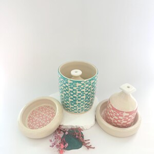 May include: Two ceramic incense holders with matching lids. The larger holder is teal with a white geometric pattern. The smaller holder is pink with a white geometric pattern. Both holders are sitting on a white surface.