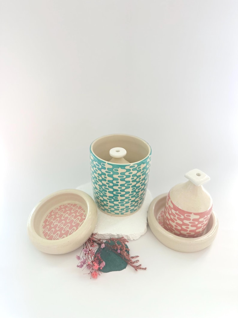May include: Two ceramic incense holders with matching lids. The larger holder is teal with a white geometric pattern. The smaller holder is pink with a white geometric pattern. Both holders are sitting on a white surface.