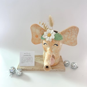 Puede incluir: Un jarrón de cerámica con forma de elefante en tonos melocotón y crema, adornado con una margarita blanca y hojas verdes. El elefante tiene una trompa y orejas grandes. El jarrón está decorado con flores secas y se apoya sobre una base de piedra, con adornos de bolas de discoteca.
