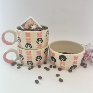 Puede incluir: Tres tazas de cerámica con un patrón rosa y blanco que presenta una silueta negra de una mujer con gafas de sol. Las tazas están apiladas una encima de la otra. Hay granos de café esparcidos alrededor de las tazas.