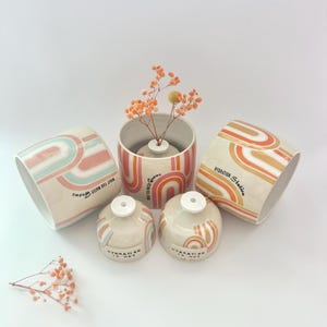 Op de afbeelding: Een verzameling keramische vazen en kleine potjes met een retro regenboogontwerp in perzik, roze en teal. De vazen hebben de tekst "What You Water Grows" en "Hydration Station". De potjes zeggen "Hydration is Hot".