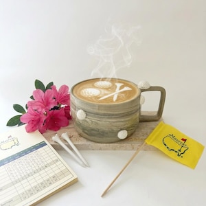 Puede incluir: Una taza de café con temática de golf con un diseño de café con leche de pelotas de golf y un tee. La taza está adornada con detalles de pelotas de golf. Una bandera de Masters, una tarjeta de puntuación, flores rosas y tees de golf completan la escena.