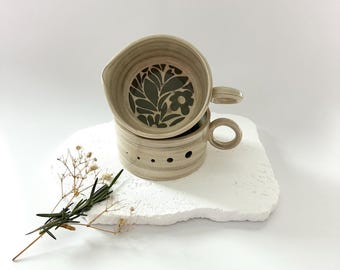 May include: Two beige ceramic mugs with handles are stacked on a white, textured surface. The top mug has a spout and a dark green floral design inside. The bottom mug has small holes around the side.