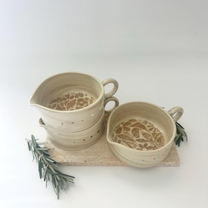 May include: A set of handmade ceramic serving bowls in a neutral beige color. Each bowl features a unique floral pattern in the base and a small handle. The bowls are stacked on a rectangular stone tray, with sprigs of rosemary.