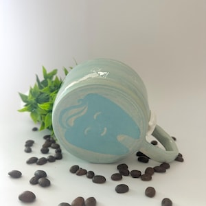 May include: A ceramic mug with a light blue and white swirl design, resting on its side. The mug is surrounded by scattered coffee beans and a small green plant. The mug's handle is white.
