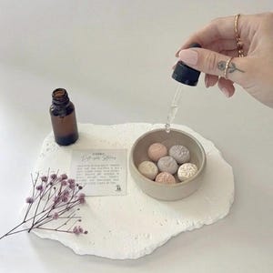 Peut inclure: Un ensemble de pierres de diffusion en céramique avec une petite bouteille marron et un compte-gouttes. L'ensemble comprend un petit bol avec des pierres décoratives roses, grises et blanches. Une brindille de fleurs violettes et une carte sont également présentes.