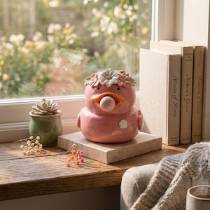 May include: A pink ceramic duck-shaped planter with a flower crown and a small white flower detail. The planter sits on a square stone block, next to a small green pot with a succulent. The scene is set on a wooden windowsill with books and a cozy blanket.