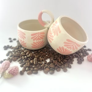 Puede incluir: Dos tazas de cerámica blanca con diseños florales rosas, sentadas sobre un lecho de granos de café.