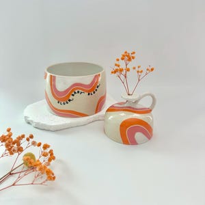 Op de afbeelding: Twee keramische vazen met een crèmekleurige basis en oranje en roze golvende strepen. De grotere vaas heeft de tekst "BLOOM BABY BLOOM". De kleinere vaas heeft een handvat en bevat oranje bloemen.
