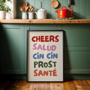 May include: A framed print with the word "CHEERS" in red, "SALUD" in purple, "CIN CIN" in blue, "PROST" in green, and "SANTÉ" in orange. The print is in a black frame and is displayed in a kitchen setting.