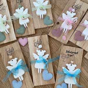 May include: Wooden angel ornaments with the text "Schutzengel to go". Each ornament features a white angel figure with ribbon, a heart, and a wooden base. The hearts are in various colors, including pink, blue, and green.